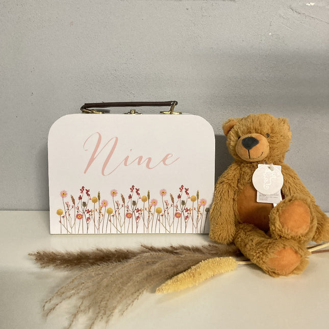 Een witte kinderkoffer met naam en bloemen erop en daarnaast is er een knuffel