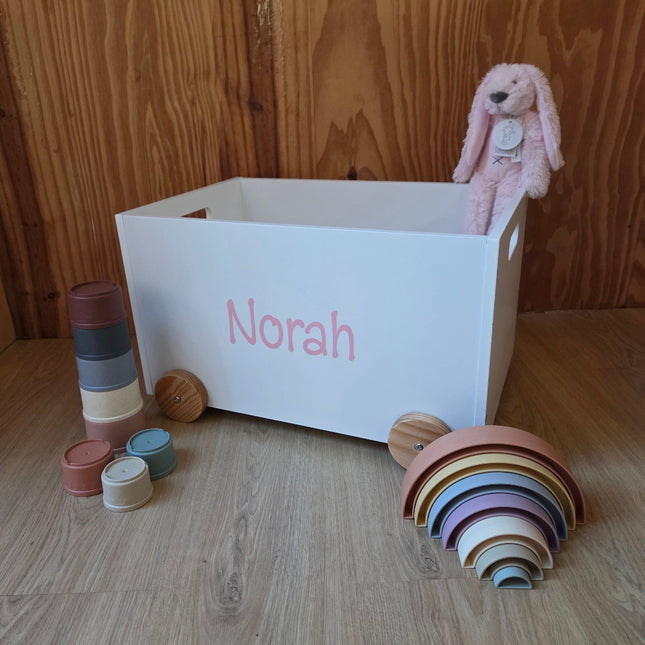 Een witte speelgoedkist op wielen met de naam van het kindje er op in pastel roze, met leuk speelgoed